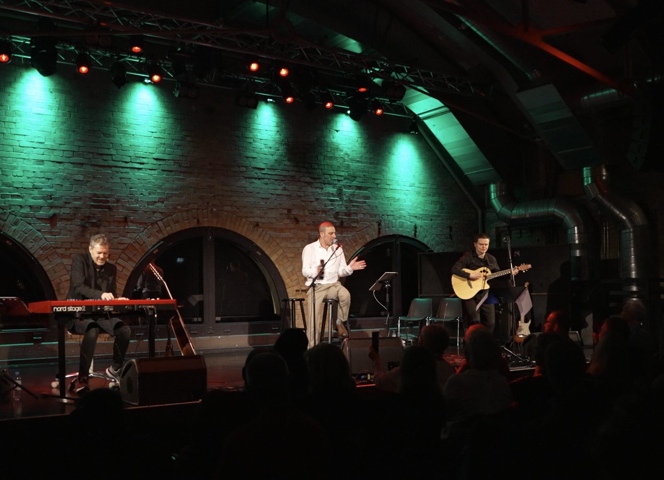 Greg Hunting am Nord-Keyboard beim Coaching-Konzert, dazu Sänger am Mikrofon und Gitarrist, grün und rot beleuchtete Bühne der Kulturbrauerei Berlin