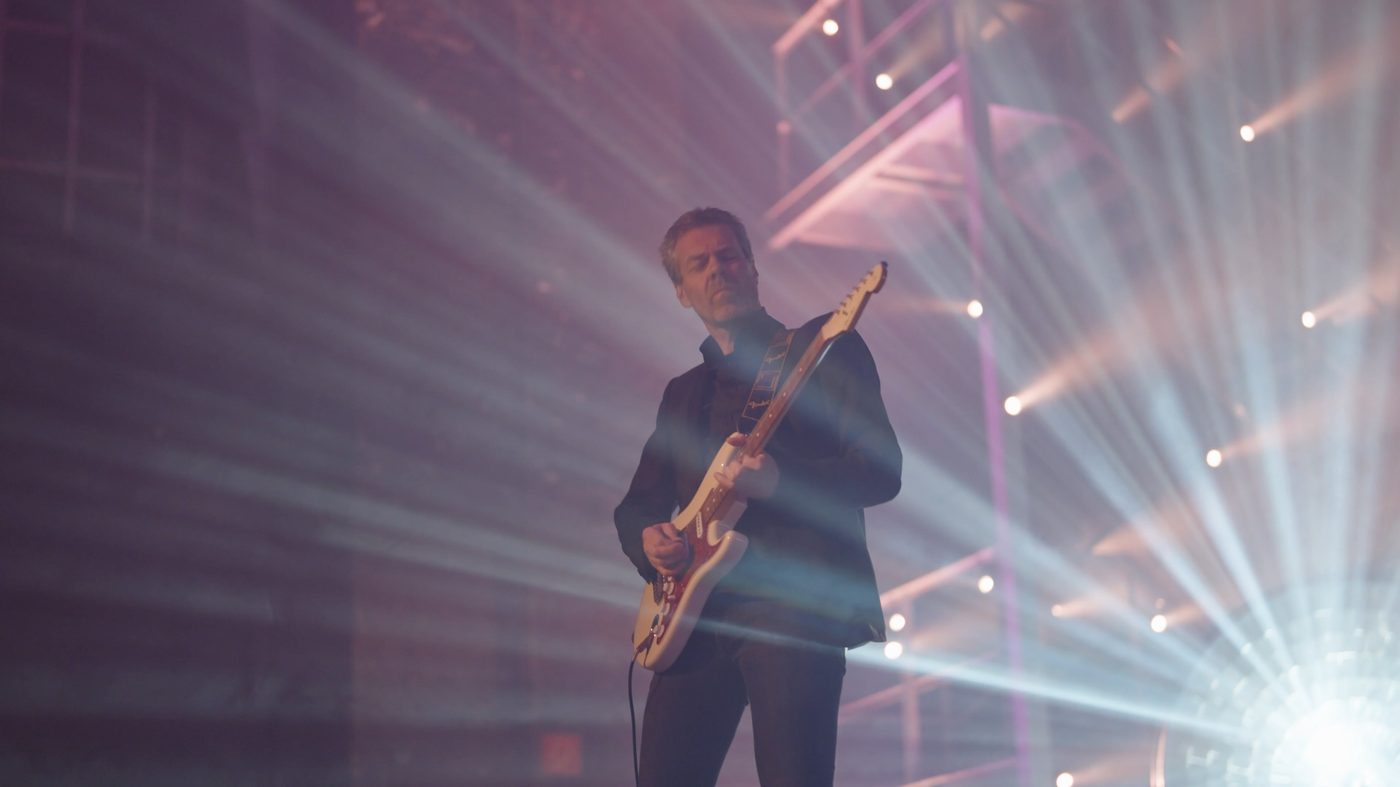 Greg Hunting steht mit Gitarre auf dunkler Bühne, hinter ihm fächern Lichtstrahlen auf
