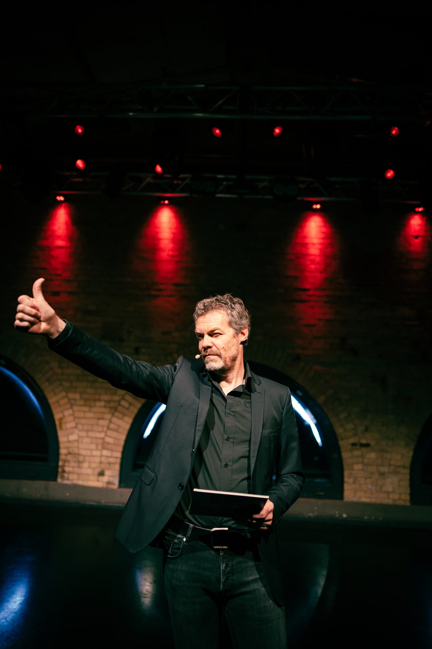 Greg Hunting auf der Bühne mit erhobenem Arm und Daumen hoch, weite Bühne im roten Licht